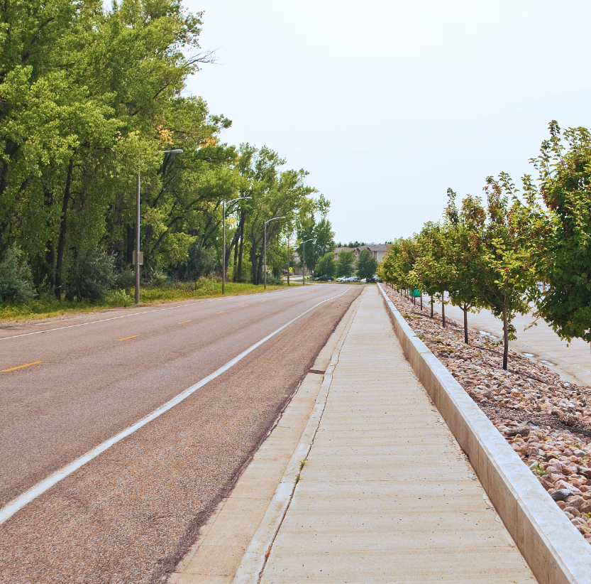 Roadway with raised sidewalk next to a slanted rock bed with newly planted trees.